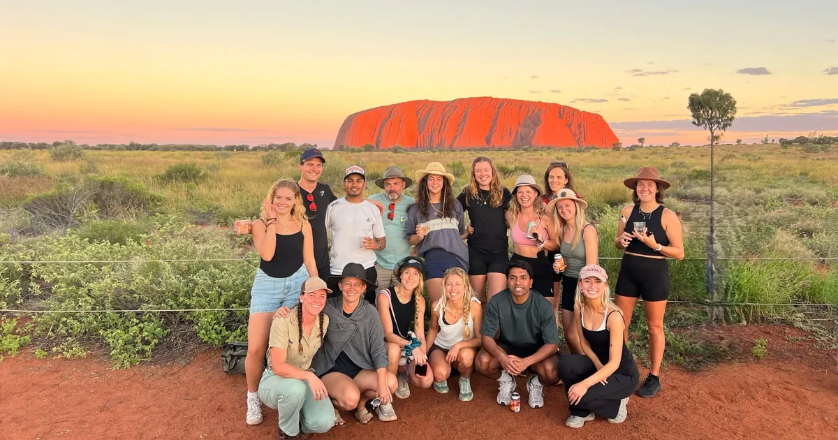 Un gruppo di un tour riunito nella terra rossa al tramonto con Uluru all'orizzonte alle spalle