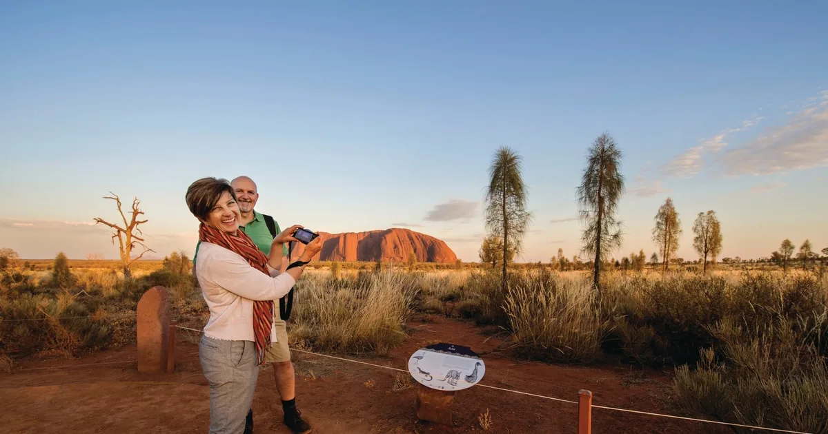 Una donna che fotografa Uluru all'alba da un'area panoramica recintata, un uomo accanto a lei e querce del deserto intorno