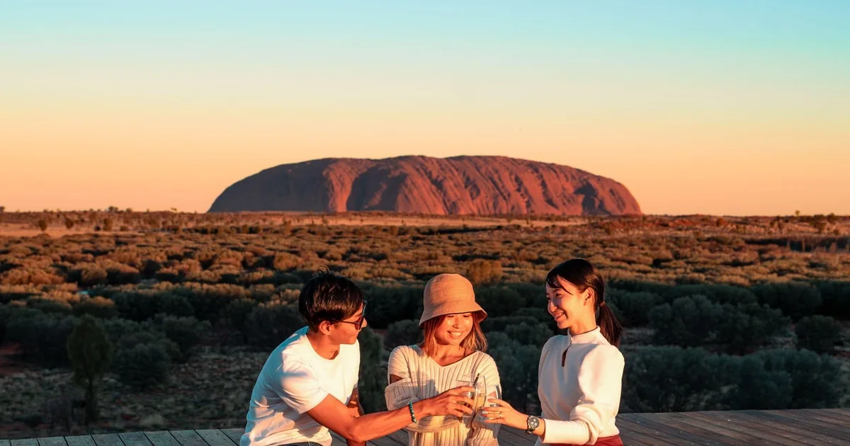Drei Menschen erheben ihre Gläser auf einer Aussichtsplattform bei Sonnenuntergang, am Horizont der Uluru