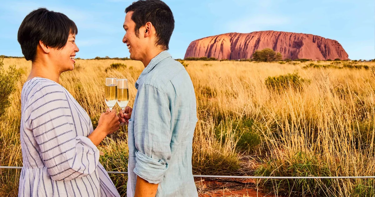Ein Paar mit Gläsern Sekt im goldenen Gras, hinter ihnen der Uluru im Abendlicht