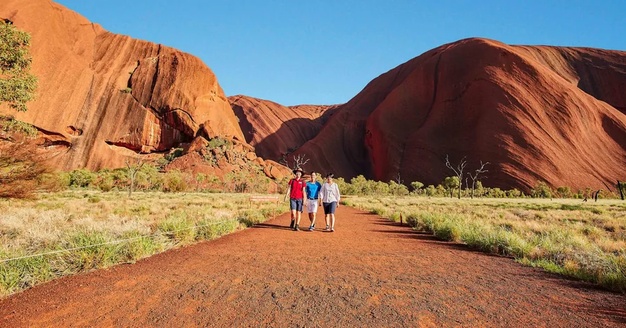 Eine kleine Wandergruppe auf einem Pfad unter der steilen, gerillten roten Wand des Uluru