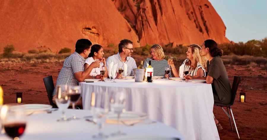 Eine Gruppe beim Essen an einem weiß gedeckten Tisch in der Wüste in der Dämmerung, am Horizont der Uluru