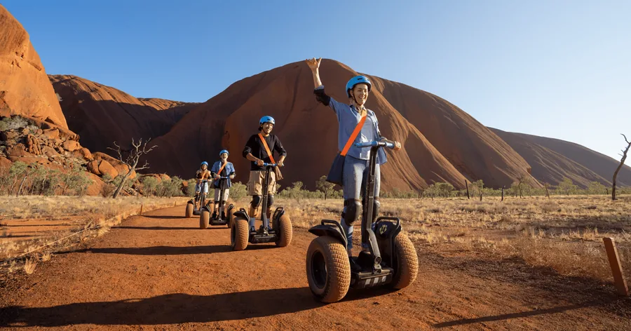 Eine Reihe von Segway-Fahrern auf einem roten Weg, hinter ihnen ragt am Morgen der Uluru auf